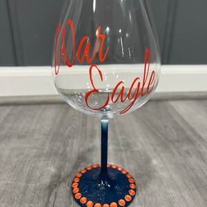 Puede incluir: Una copa de vino de vidrio transparente con un tallo y una base azul marino. La base está decorada con un círculo de puntos naranjas. La copa tiene las palabras "War Eagle" escritas en rojo en el lateral.