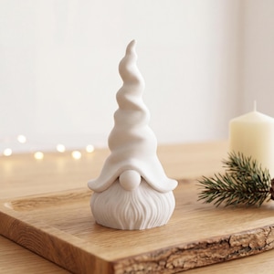 Puede incluir: Una figura de gnomo de cerámica blanca mate con un sombrero puntiagudo en espiral y una barba texturizada. El gnomo está colocado sobre una bandeja de madera junto a una vela de pilar color crema, una pequeña rama de pino y una piña.