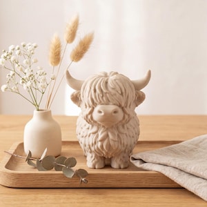 Op de afbeelding: Een beige Schotse Hooglander-figuur met gestructureerde vacht en gebogen hoorns, geplaatst op een houten dienblad naast een klein wit vaasje met droogbloemen en een takje eucalyptus, vergezeld van een gevouwen lichtbruin linnen servet.