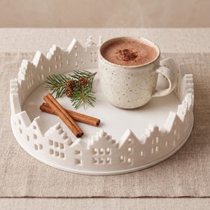 Könnte beinhalten: Ein rundes, weißes Keramiktablett mit einem dekorativen Rand aus kleinen Häusersilhouetten. Auf dem Tablett stehen eine gesprenkelte beige Tasse mit einem braunen Getränk, ein kleiner grüner Tannenzweig und zwei braune Zimtstangen.