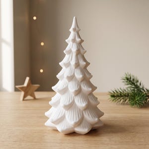 Op de afbeelding: Een wit, kegelvormig sculpturaal object in de vorm van een gestileerde kerstboom met gelaagde, bloembladachtige takken. Het oppervlak heeft verticale ribbels en een gladde, matte afwerking, staand als een decoratief tafelornament.