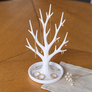 Può includere: Un espositore per gioielli bianco a forma di albero senza foglie con molteplici rami per appendere collane e orecchini. La base è un vassoio circolare che contiene diversi anelli color oro e una sottile catenina d'oro come supporto.