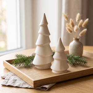 Könnte beinhalten: Zwei glatte, mattweiße Keramik-Baumfiguren in gestufter, konischer Form. Der größere Baum hat fünf ausgeprägte Ebenen, während der kleinere Baum drei Ebenen aufweist. Beide haben eine spitze Oberseite und einen flachen, runden Sockel.