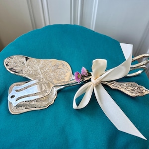 May include: Silver serving set with ornate floral detailing, including a cake server and a pie server. The set is tied together with a cream-colored ribbon and small pink flowers. The set is displayed on a teal surface.
