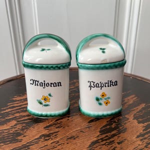Può includere: Due barattoli per spezie in ceramica bianca con coperchi verdi e disegni floreali. I barattoli sono etichettati "Majoran" e "Paprika".