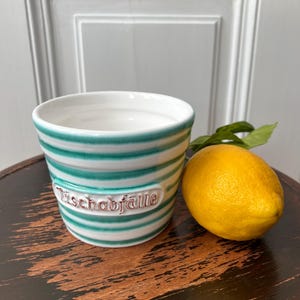 Puede incluir: Un recipiente de cerámica blanca con rayas verdes y la palabra "Tisch Abfall" en letras rosas. El recipiente está sobre una superficie de madera junto a un limón amarillo.