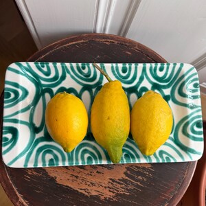 Può includere: Tre limoni giallo brillante sono disposti su un vassoio rettangolare in ceramica. Il vassoio presenta uno sfondo bianco con un motivo a spirale turchese. I limoni sono disposti orizzontalmente. Il vassoio poggia su una superficie di legno scuro.