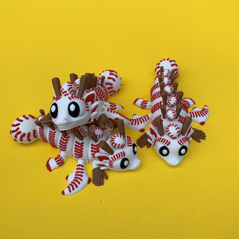 May include: Three whimsical axolotl figurines, each creatively designed with baseball-themed elements. The creatures feature white bodies with red stitching, brown appendages, and large, expressive eyes, set against a vibrant yellow background.
