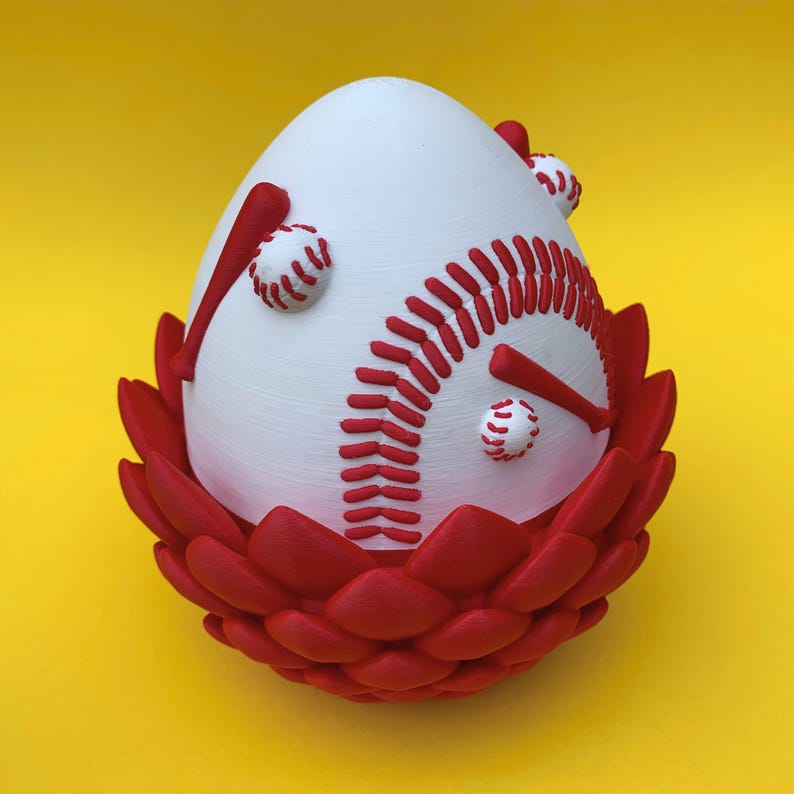 May include: A baseball-themed decorative egg, white with red stitching and baseball bat accents. The egg sits in a red, petal-like base against a yellow background. A unique sports-themed decorative item.