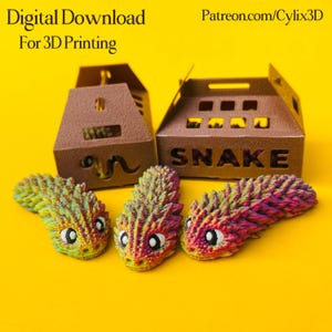 Op de afbeelding: Een afbeelding van 3D-geprinte slangenfiguren in groen, geel en roze, samen met bruine kartonnen dozen. Op de dozen staat "SNAKE" en "an". De tekst "Digital Download For 3D Printing" is zichtbaar bovenaan.