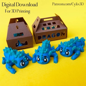 May include: Three blue 3D-printed dragon figurines with crystal-like spines and large eyes are displayed on a yellow surface. Two brown boxes with cut-out designs and the word "DRAGON" are in the background. Text at the top reads "Digital Download For 3D Printing" and "Patreon.com/Cylix3D".