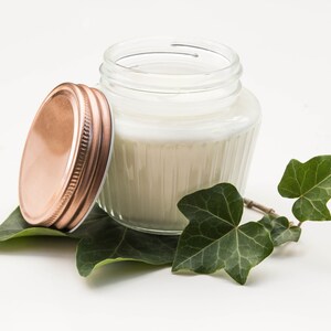 Könnte beinhalten: Ein klares Glas mit weißer Creme und einem kupferfarbenen Deckel daneben. Das Glas steht auf grünen Efeublättern, was eine natürliche Ästhetik erzeugt. Die Creme wirkt glatt und dick. Wahrscheinlich ein Kosmetikprodukt.