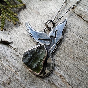 Peut inclure: Un collier pendentif en argent présentant un motif découpé d'oiseau en vol aux ailes déployées. Le pendentif est serti d'une pierre brute vert olive foncé à la base et est suspendu à une fine chaîne en argent avec de petites perles métalliques.