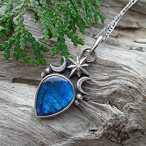 May include: A silver pendant necklace featuring a teardrop-shaped, blue labradorite stone. The pendant is adorned with a star and crescent moon design. The necklace is displayed on a piece of weathered wood, with green foliage in the background.