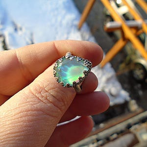 Pode incluir: Um anel de prata com uma grande opala em forma de pêra com tons verdes e azuis iridescentes. A opala é definida em uma moldura detalhada e ornamentada. O anel é segurado contra um fundo desfocado ao ar livre.