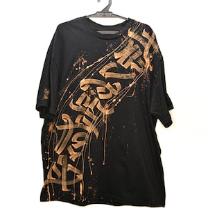 Puede incluir: Una camiseta negra de manga corta con un diseño caligráfico diagonal de color claro en la parte delantera. El gráfico incluye efectos de pintura salpicada y goteante en un tono bronceado que se extienden por la superficie de la tela.