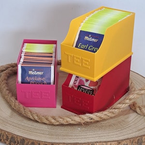 Könnte beinhalten: Drei Teebeutel-Organizer, einer gelb, einer rot und einer pink, jeweils mit dem Wort "TEE" auf der Vorderseite geprägt. Der gelbe Organizer hat den Text "Meßmer Earl Grey" auf der Vorderseite, der pinke Organizer hat den Text "Meßmer Aprikose-Pfirsich" auf der Vorderseite und der rote Organizer hat ein Blumenmuster mit dem Text "A BAG OF OUR SPECIAL" auf der Vorderseite.