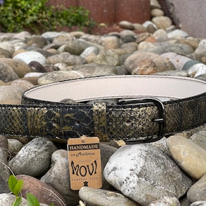 May include: A black leather belt with a gold embossed pattern. The belt has a black metal buckle and a brown tag that reads "Handmade in Mallorca, Since 1979, MOUI, Leather 100%".