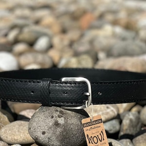 Könnte beinhalten: Ein schwarzer Ledergürtel mit einer silbernen Schnalle. Der Gürtel ist mit weißem Garn vernäht. Der Gürtel ist handgefertigt in Mallorca, Spanien. Das Etikett lautet "MOVI seit 1978, Leder 100%"