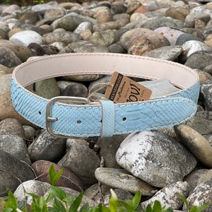 May include: A light blue leather belt with a snakeskin pattern and a silver buckle. The belt is handmade in Italy and has white stitching detail. The belt is lying on a bed of small grey stones.