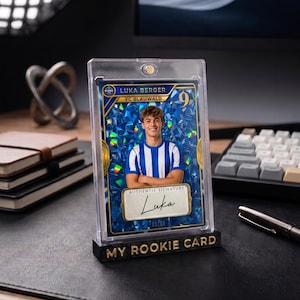 May include: A rectangular trading card featuring a portrait of a soccer player in a blue and white striped jersey, set against a blue holographic background. The card displays the text Luka Berger, FC Blauwald, and an authentic signature label.
