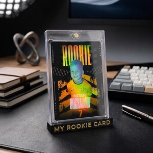 May include: A rectangular trading card featuring a photograph of a child in a yellow sports jersey, encased in a clear magnetic acrylic holder. The card displays the text ROOKIE, NOAH BOCKMANN, and the number 7. A black base with the text MY ROOKIE CARD.