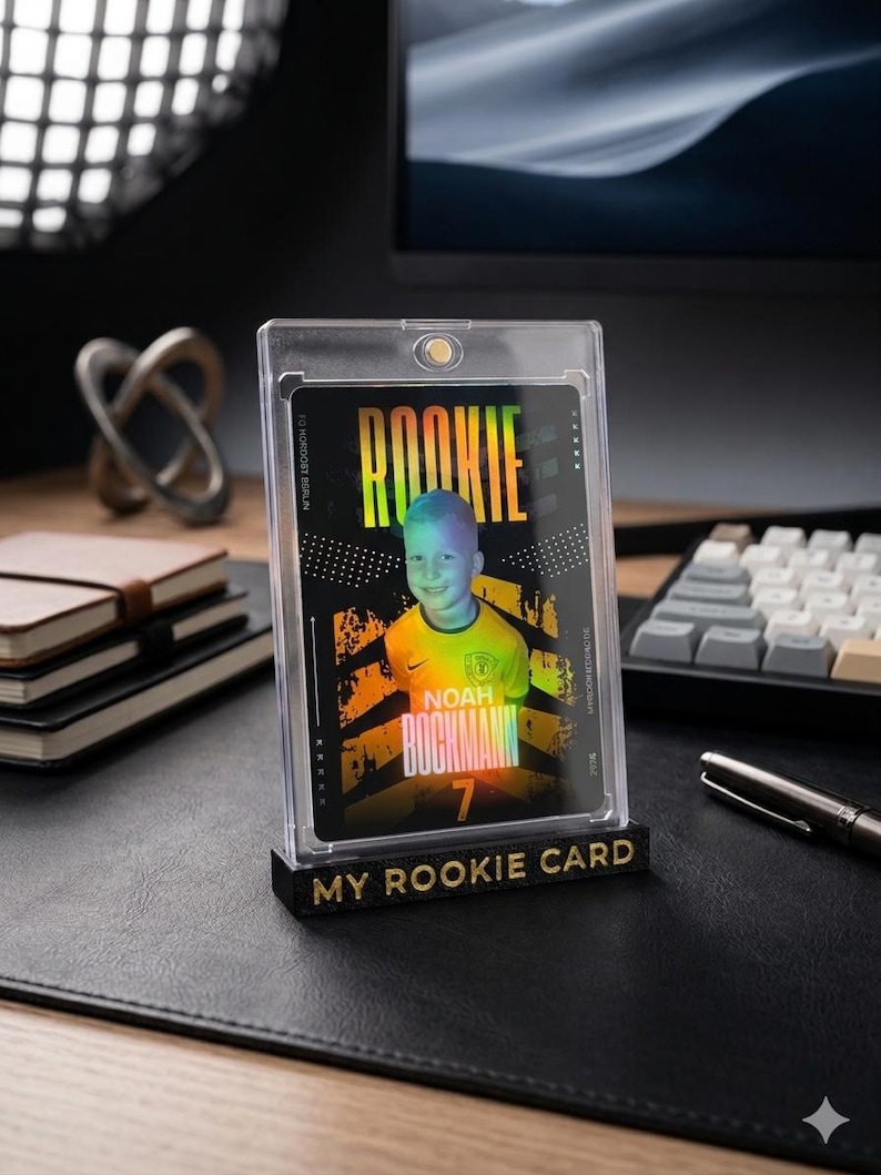 May include: A rectangular trading card featuring a photograph of a child in a yellow sports jersey, encased in a clear magnetic acrylic holder. The card displays the text ROOKIE, NOAH BOCKMANN, and the number 7. A black base with the text MY ROOKIE CARD.