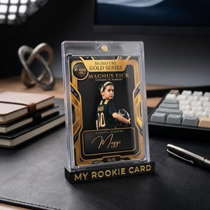 May include: A collectible sports trading card encased in a clear magnetic holder, featuring a young soccer player named Magnus Eick. The card has gold accents, the text "Signature Gold Series," "My Rookie Card," and a signature labeled "Maggi."