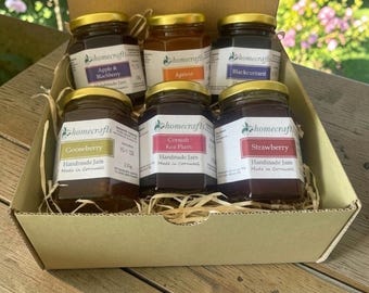 May include: A cardboard gift box containing six glass jars of Homecrafts handmade jam. The flavours include Apple & Blackberry, Apricot, Blackcurrant, Gooseberry, Cornish Kea Plum, and Strawberry, each with a gold lid and a white label.