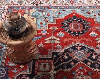 May include: A detailed shot of a red, blue, and cream-colored rug with an intricate geometric pattern. A small, woven stool holds a dark glass vase. The rug's design features floral and geometric elements, creating a rich, textured appearance.