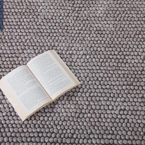May include: A close-up of a gray, textured rug made of small, round balls. A book is lying on top of the rug.