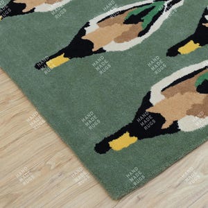 Könnte beinhalten: Grüner Teppich mit einem sich wiederholenden Entenmuster. Die Enten sind mit weißen, braunen, schwarzen, grünen und gelben Details dargestellt. Die Worte "HAND MADE RUGS" sind auf dem Teppich aufgedruckt.