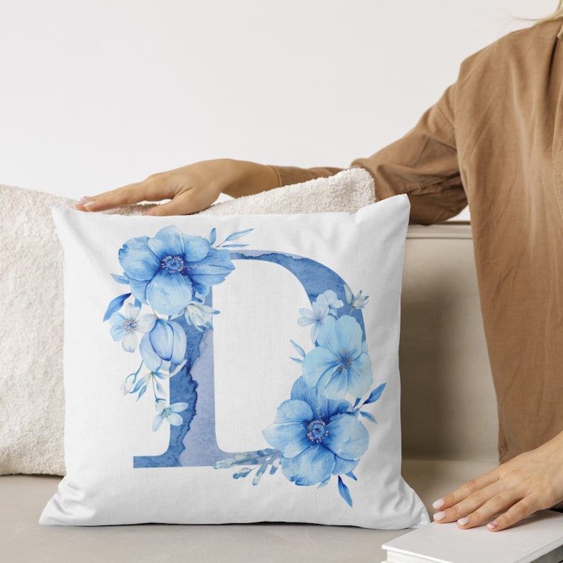 May include: White pillow with a blue watercolor letter D surrounded by blue flowers. The letter D is stylized with a watercolor effect.