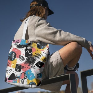 Puede incluir: Una bolsa de tela blanca con un patr&oacute;n geom&eacute;trico colorido. El patr&oacute;n incluye pinceladas negras, formas amarillas, rosas, azules y blancas. La bolsa tiene asas negras.