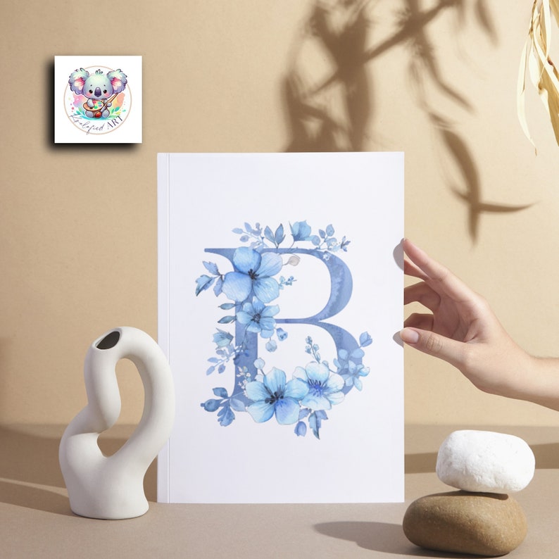 May include: A watercolor print of the letter B surrounded by blue flowers. The print is on white paper and has a white border.