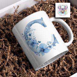 May include: White ceramic mug with a blue watercolor letter C surrounded by blue flowers. The mug is sitting in brown packing peanuts.