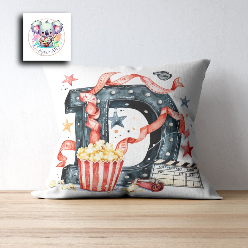 May include: White pillow with a red, white, and blue illustration of a movie themed letter D. The letter is made of a black metal with light bulbs. There is a red ribbon, popcorn in a red and white striped bucket, and a movie clapperboard with the text "COVENTREY" on it.