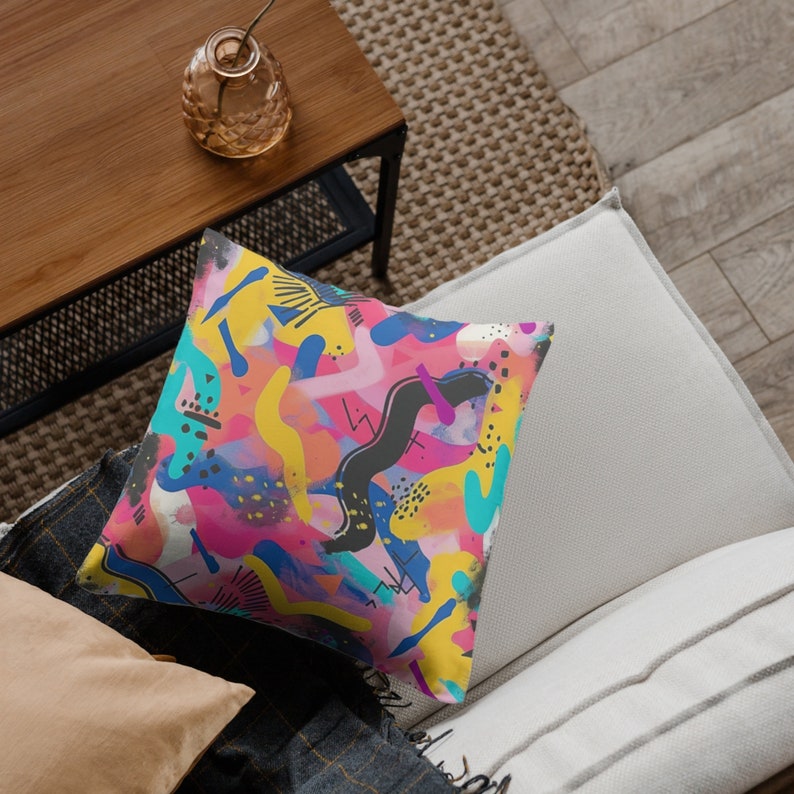 Puede incluir: Una almohada de tiro colorida con un patr&oacute;n geom&eacute;trico abstracto en tonos de rosa, amarillo, azul y negro. La almohada est&aacute; en un sof&aacute; blanco.