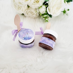 Peut inclure: Deux petits pots en verre avec couvercles, remplis de pâte à tartiner au chocolat, décorés d'une étiquette violette et blanche représentant un personnage de dessin animé et le texte "Shana 4 anos". Chaque pot est muni d'une cuillère en bois attachée avec un ruban violet.
