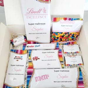 Peut inclure: Un coffret cadeau rempli de divers bonbons et chocolats, dont Lindt Excellence, Haribo, Kinder, Kit Kat et M&M's. L'emballage présente des motifs de crayons colorés et le texte "Super maîtresse Sophie".