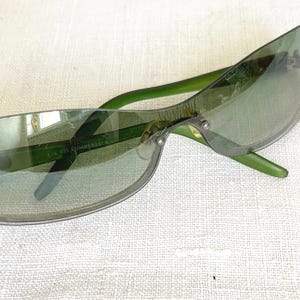 Puede incluir: Gafas de sol con montura verde y lentes tintadas. Las gafas tienen forma rectangular y un diseño elegante. Las patillas son de color verde vibrante y las lentes tienen un tinte degradado. Las gafas descansan sobre una superficie blanca.