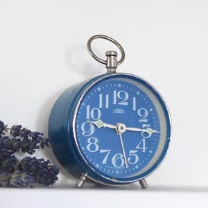May include: A round analog alarm clock with a deep blue face, white Arabic numerals, and silver-toned hands. The clock features a metallic silver casing, a top ring handle, and two small feet. The brand name Prim is printed on the clock face.