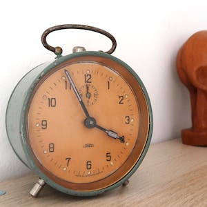 May include: Vintage green and tan alarm clock with a curved handle. The clock face has black numerals and hands, with a small dial at the top. The clock is set on a wooden surface.