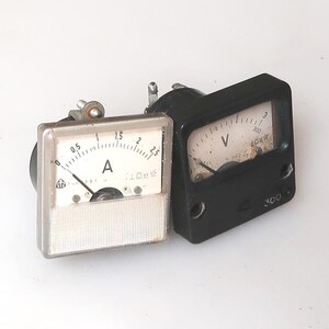 Puede incluir: Dos medidores eléctricos antiguos. Uno es un voltímetro negro con una esfera blanca y una aguja que apunta hacia la izquierda. El otro es un amperímetro con esfera transparente, aguja negra y la letra "A".