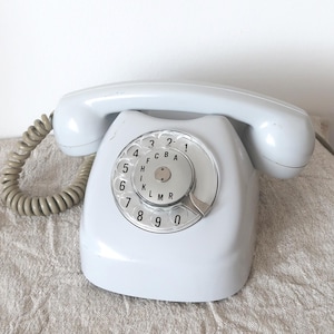 Op de afbeelding: Een vintage lichtblauwe draaischijftelefoon met bijpassende handset en spiraalsnoer. De draaischijf heeft cijfers en letters. De telefoon staat op een getextureerd, lichtgekleurd oppervlak.