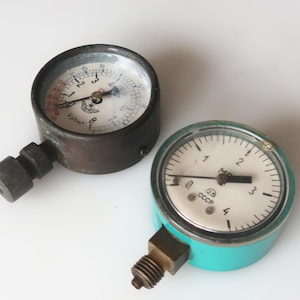 Può includere: Due manometri vintage. Uno è in metallo scuro con quadrante bianco e segni rossi. L'altro è turchese con quadrante bianco e segni neri. Entrambi i manometri hanno un quadrante circolare e un raccordo filettato.
