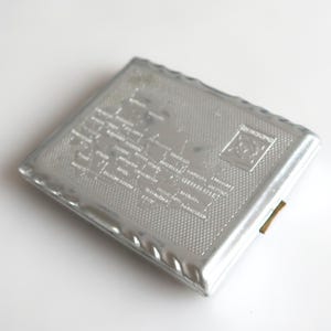 May include: A silver metal cigarette case with a textured surface and a small gold hinge. The case features a design of a map with various cities and towns printed on it.