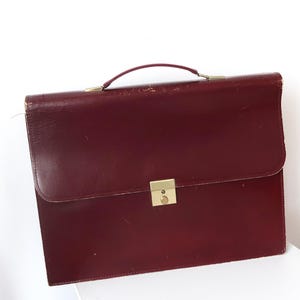 May include: A dark burgundy leather briefcase featuring a top handle and a rectangular gold-tone metal clasp closure. The structured bag has visible signs of wear along the top edges and a smooth, matte finish on the front flap surface.