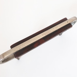 Puede incluir: Un tirador de cajón con una pieza central rectangular de patrón de carey marrón oscuro, enmarcado por una estructura de metal plateado cepillado con extremos redondeados y dos postes de montaje en la parte inferior del marco.