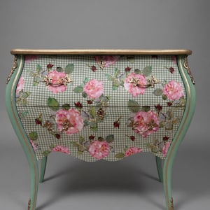 May include: A two-drawer chest of drawers with a floral design. The chest has a light green frame with gold accents and curved legs. The drawers are covered in a green and white checkered pattern with pink roses.